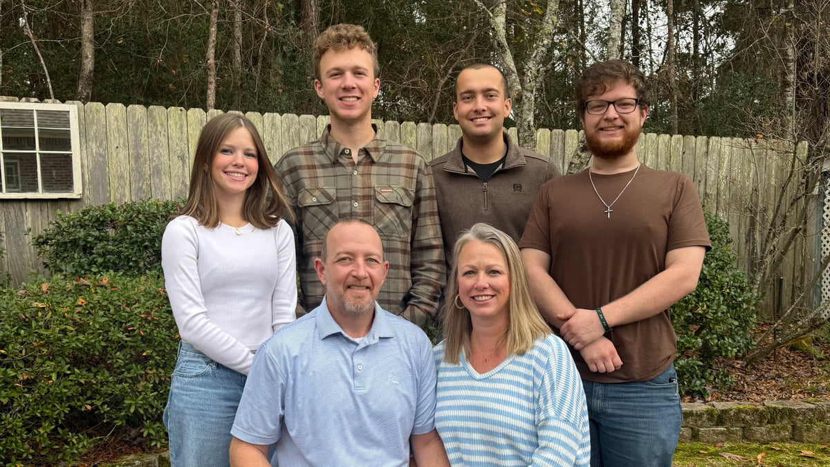 A family of five posing together outdoors in a backyard setting with trees and a wooden fence behind them.